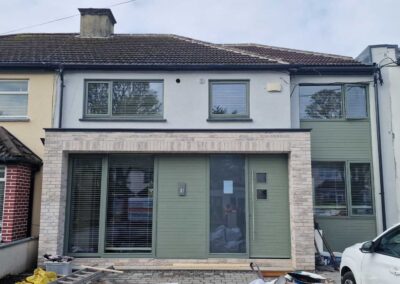 Renovated house facade with light brick cladding, large windows with blinds, and modern green front door